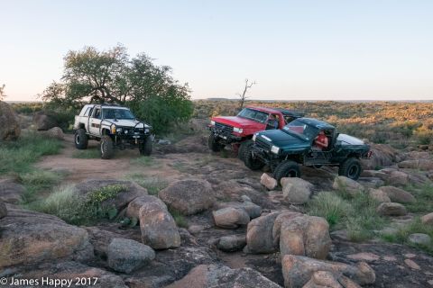 1989 Ford Bronco - Modified trucks traverse a rocky terrain, with a white SUV on the left and red and green trucks on the right.