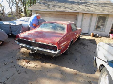 1968 Mercury Cougar - A man inspects a red 1968 Mercury Cougar parked in a driveway with other vehicles and a garage in the background.