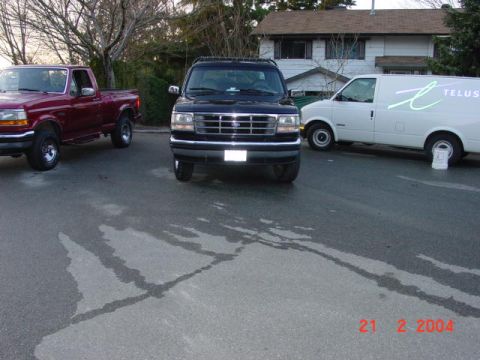 1995 Ford Bronco - A black 1995 Ford Bronco is parked on wet asphalt, with a red pickup truck and a white van nearby.