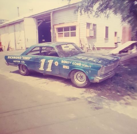 1973 Ford Mustang - A teal 1965 Ford Galaxie race car, number 11, is parked in front of a garage.
