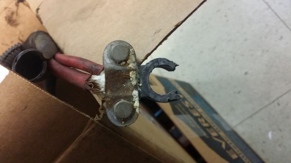 Corroded battery cable terminal with red insulated wires and a broken black rubber clamp, inside a cardboard box