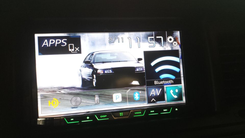 Car stereo touchscreen displaying a black sedan drifting, time 2/11 11:57, and icons for Apps, Bluetooth, AV.