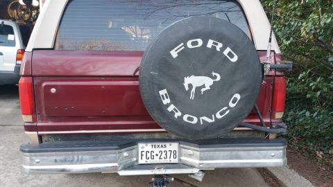 1996 Ford Bronco - The rear view of a maroon Ford Bronco with a black spare tire cover featuring a white horse logo and 'FORD BRONCO' text. A Texas license pl…