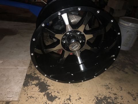 1995 Ford F-150 - A close-up, angled view of a black aftermarket wheel with chrome accents and a silver center cap. The wheel is resting on a concrete floor.