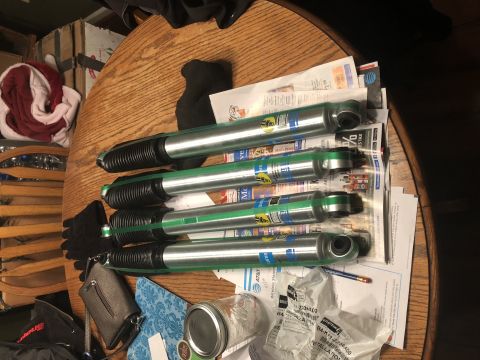 1995 Ford F-150 - Four Bilstein shock absorbers with green bands and black bellows are laid out on a wooden table, partially covering some papers and other i…