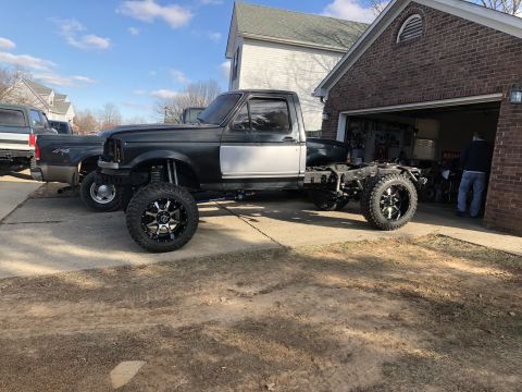 1995 Ford F-150 - A lifted, dark gray Ford F-150 pickup truck chassis with large aftermarket wheels is parked in a driveway next to a brick garage. Another p…