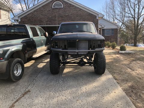 1995 Ford F-150 - The front view of a lifted, dark-colored pickup truck with its grille and headlights removed, parked on a concrete driveway in front of a b…