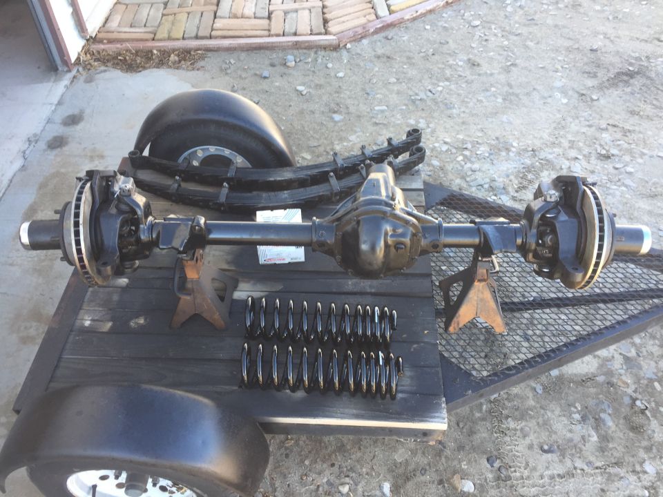 Front axle assembly with disc brakes, leaf springs, coil springs, tire, and fender on a trailer.