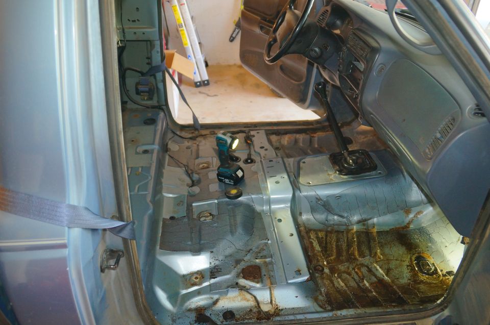 Interior of a vehicle with floor removed, showing rust and a cordless drill.
