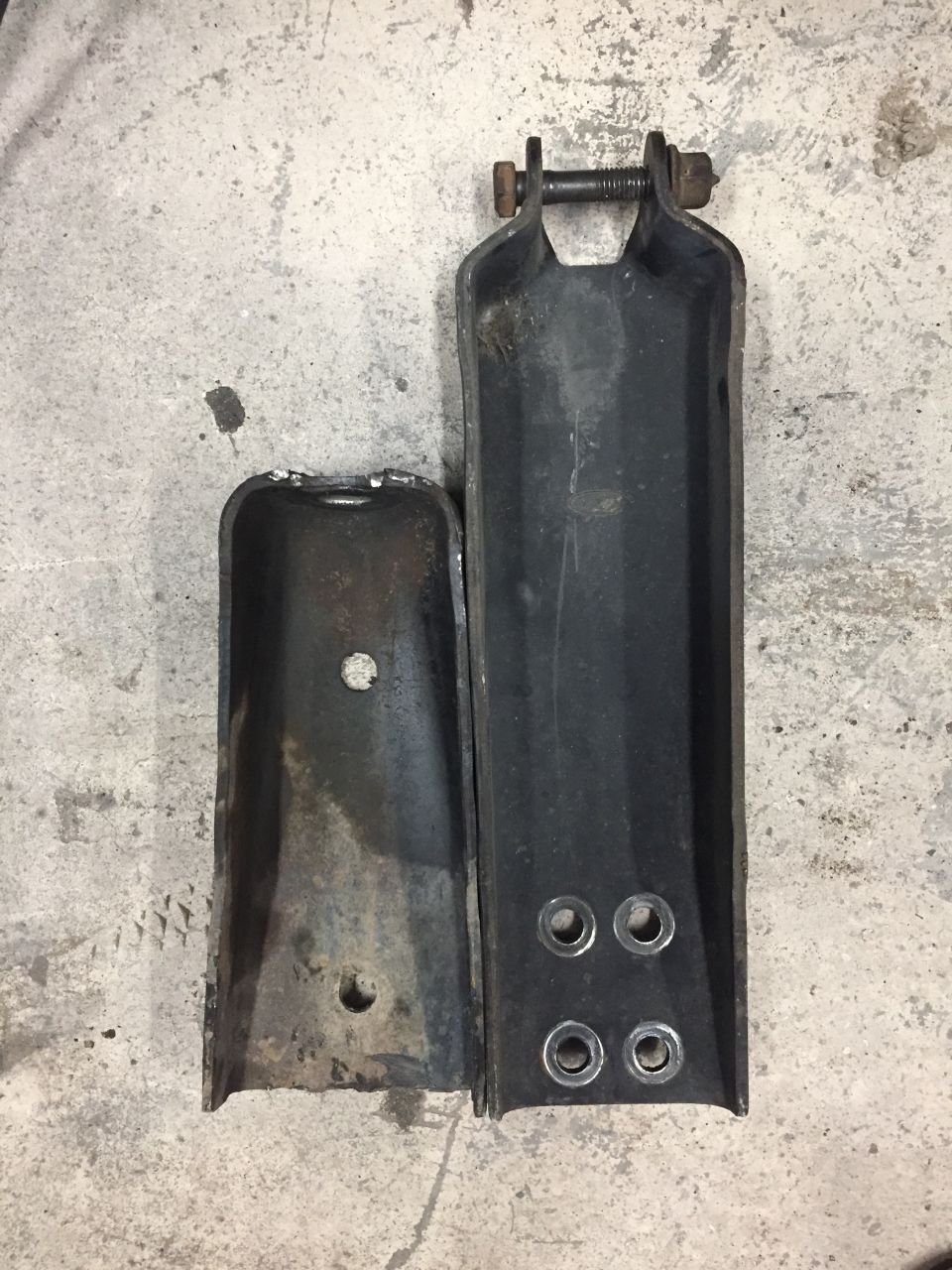 Two metal brackets on a concrete floor, one rusted and shorter, the other black and longer with grommeted holes.