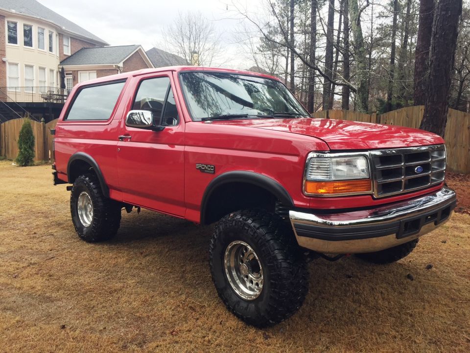 Red 1994 Ford Bronco parked on grass with a house and trees in the background.