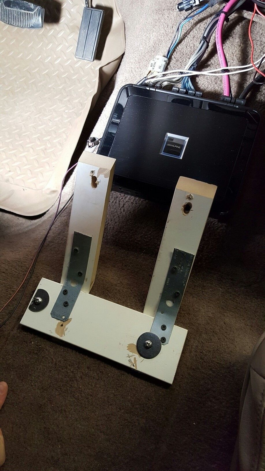 Car interior with tan carpet, pedals, and a black amplifier mounted on the floor with a wooden bracket