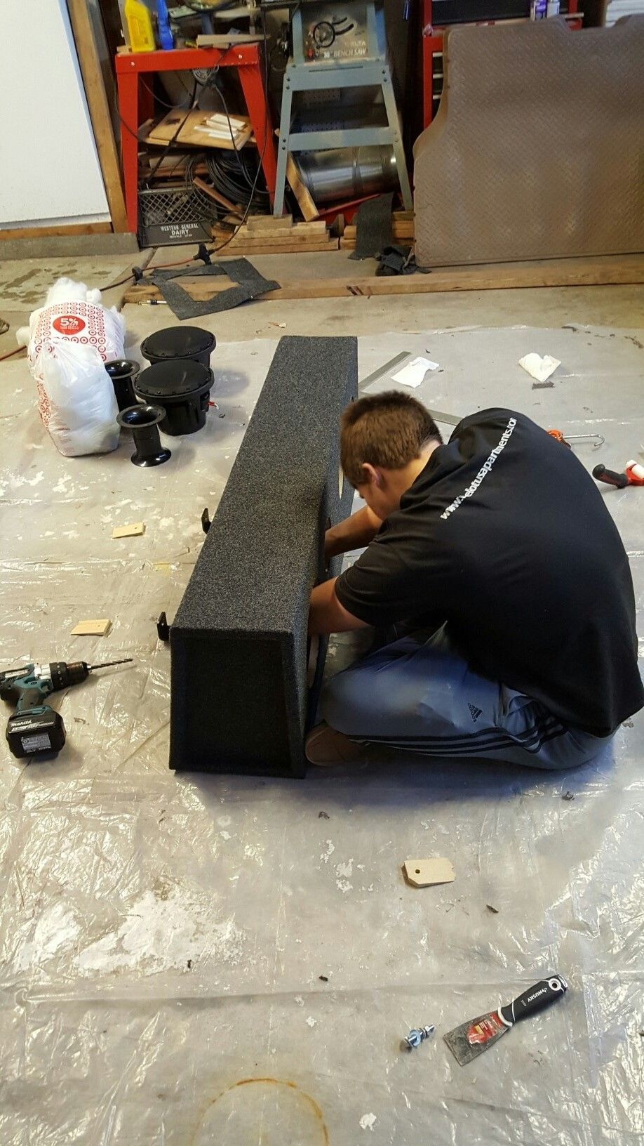 Person working on a carpeted speaker enclosure with tools and speakers nearby