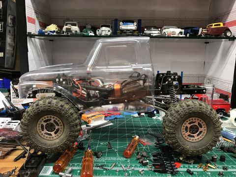 1989 Ford Bronco - An RC car chassis with a clear body and large tires sits on a workbench surrounded by tools and parts.
