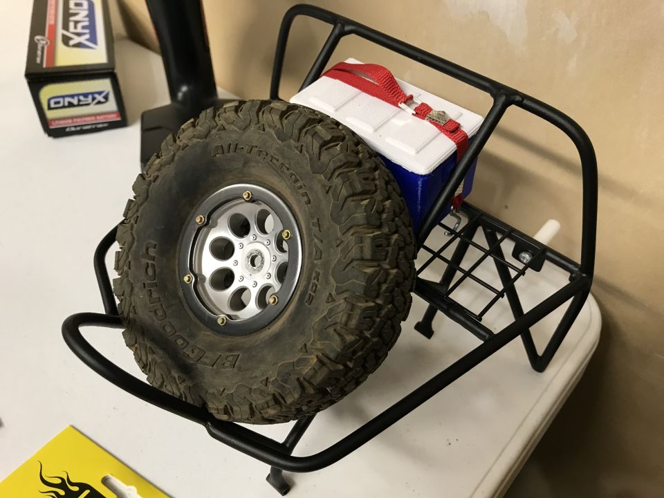Spare tire mounted on a black metal rack with a blue and white cooler secured by a red strap