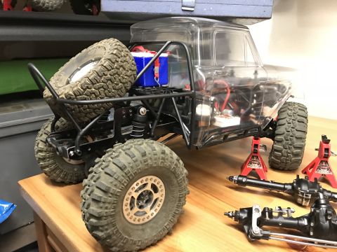 1989 Ford Bronco - RC rock crawler chassis with a clear body, spare tire mounted on the rear, and additional axles displayed on red jack stands.