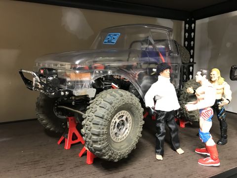 1989 Ford Bronco - A toy truck with a clear body and large tires is displayed on red jack stands, with action figures nearby.