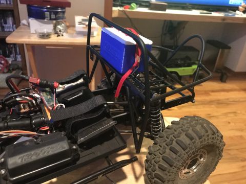 1989 Ford Bronco - An RC crawler chassis is shown with a blue cooler secured to its roll cage, with another green RC vehicle blurred in the background.