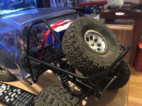 1989 Ford Bronco - The rear section of a blue RC rock crawler is shown, featuring a roll cage, a spare tire mounted on the back, and a blue battery box secure…