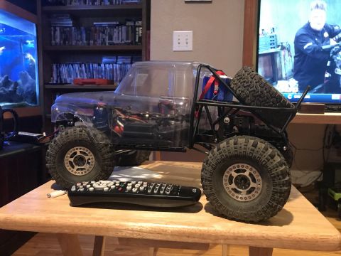 1989 Ford Bronco - A clear-bodied remote-controlled rock crawler truck sits on a wooden table in front of a bookshelf and a television. A remote control rests…