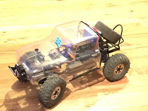 1989 Ford Bronco - A clear-bodied remote-controlled rock crawler truck with large off-road tires sits on a wooden floor. The truck features a roll cage and a…