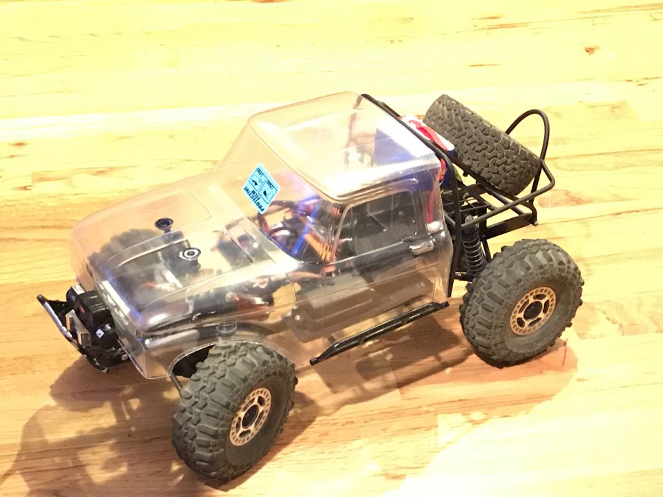 Clear-bodied RC rock crawler truck with large tires and rear roll cage on wooden floor.