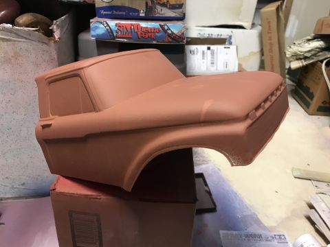 1989 Ford Bronco - A close-up, side view of a red, unpainted model truck body, likely for a radio-controlled car, resting on a cardboard box. The model appear…