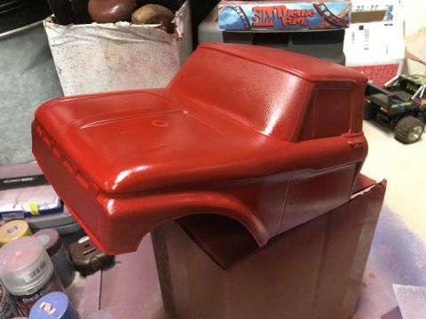 1989 Ford Bronco - A bright red, unpainted truck body is shown from a three-quarter front angle, resting on a cardboard box. Several cans of spray paint are v…