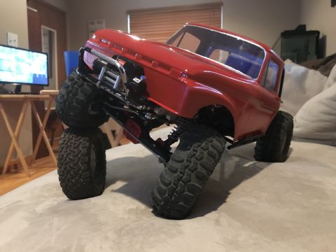 1989 Ford Bronco - A red remote-controlled rock crawler truck is shown at an extreme angle, with its front wheels stacked on top of each other. The truck is o…