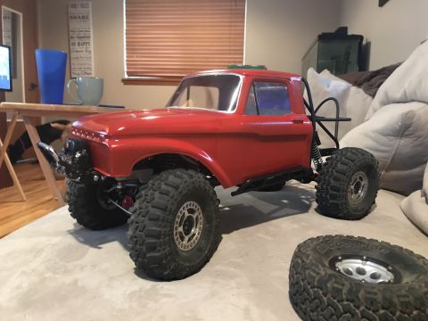 1989 Ford Bronco - A red RC rock crawler truck with large off-road tires sits on a couch. A spare tire is placed in front of the truck.