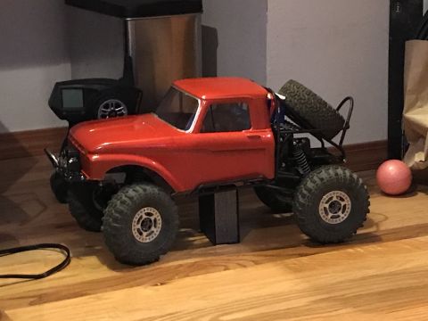 1989 Ford Bronco - A red RC rock crawler truck with large off-road tires and a spare tire mounted on the back is shown from a side angle on a wooden floor. A…