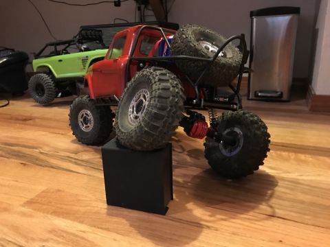 1989 Ford Bronco - A red RC rock crawler truck is displayed on a black block, with a green RC truck in the background. The red truck features large, knobby ti…