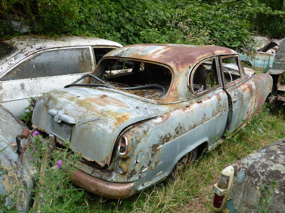 Abandoned, rusted light blue sedan with broken windows in overgrown vegetation.