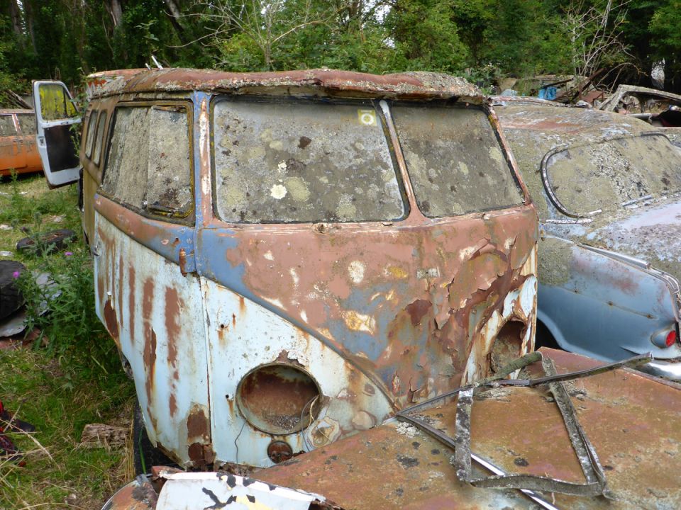 Heavily rusted vintage van with moss-covered windows in an overgrown outdoor setting.