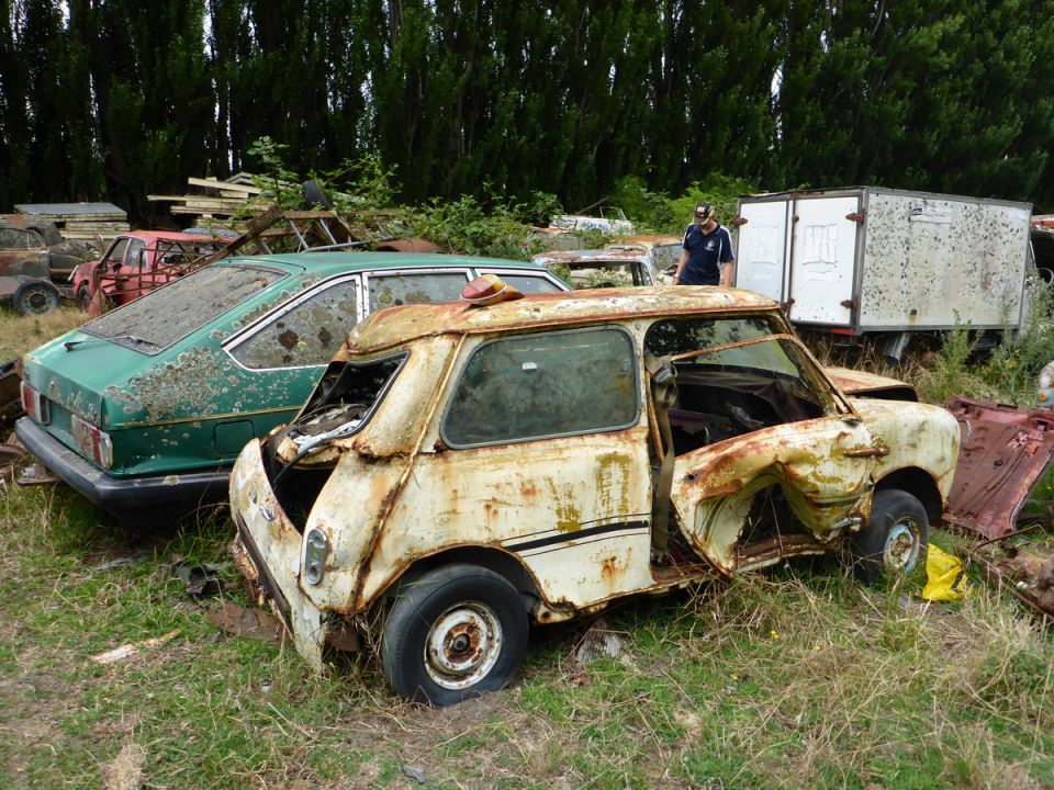 Two rusted vintage cars, a white Mini Cooper and a green sedan, in a junkyard.