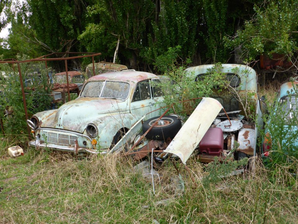 Abandoned light blue sedan in a field of weeds with other rusted vehicles.