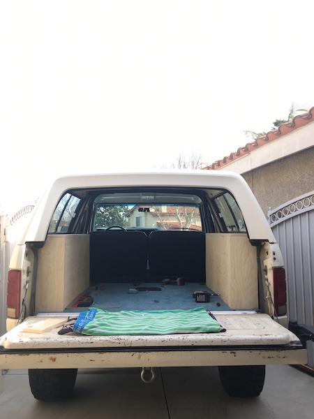 1981 Ford Bronco - Rear view of a white 1981 Ford Bronco with the tailgate down, showing a custom wooden interior paneling and blue carpeted floor. A green an…