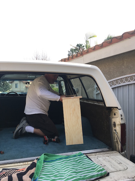 1981 Ford Bronco - A man kneels in the cargo area of a white SUV, working on a wooden box. He is holding a piece of wood and appears to be attaching it to the…