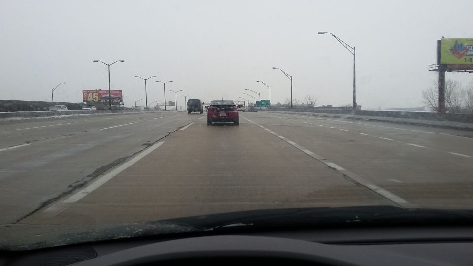 Red sedan driving on a wet highway in overcast weather.