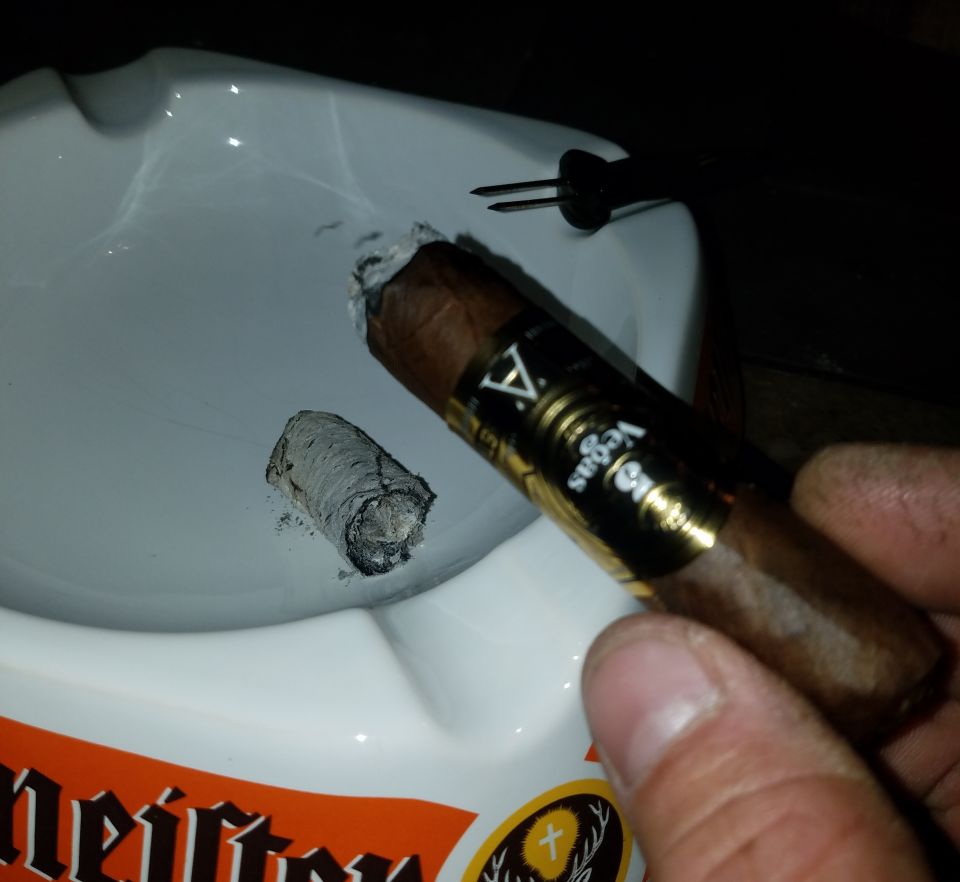 Hand holding a cigar over an ashtray with a cigar stub and partial branding.