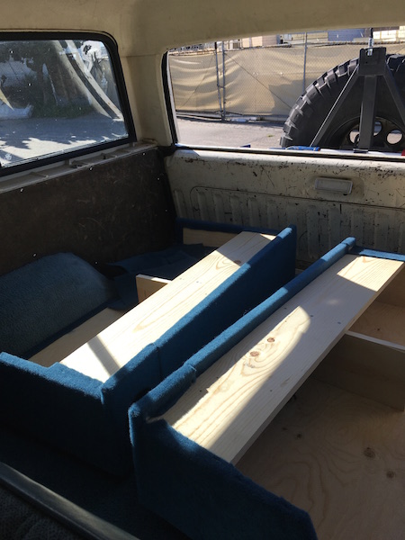 1981 Ford Bronco - The interior of a vehicle shows custom wooden platforms and blue carpeted seating, likely for a camper conversion. A large spare tire is vi…