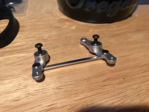 1989 Ford Bronco - A close-up shot shows a silver metal linkage assembly with two arms connected by a rod. Each arm has a screw and a bearing, and the assembl…