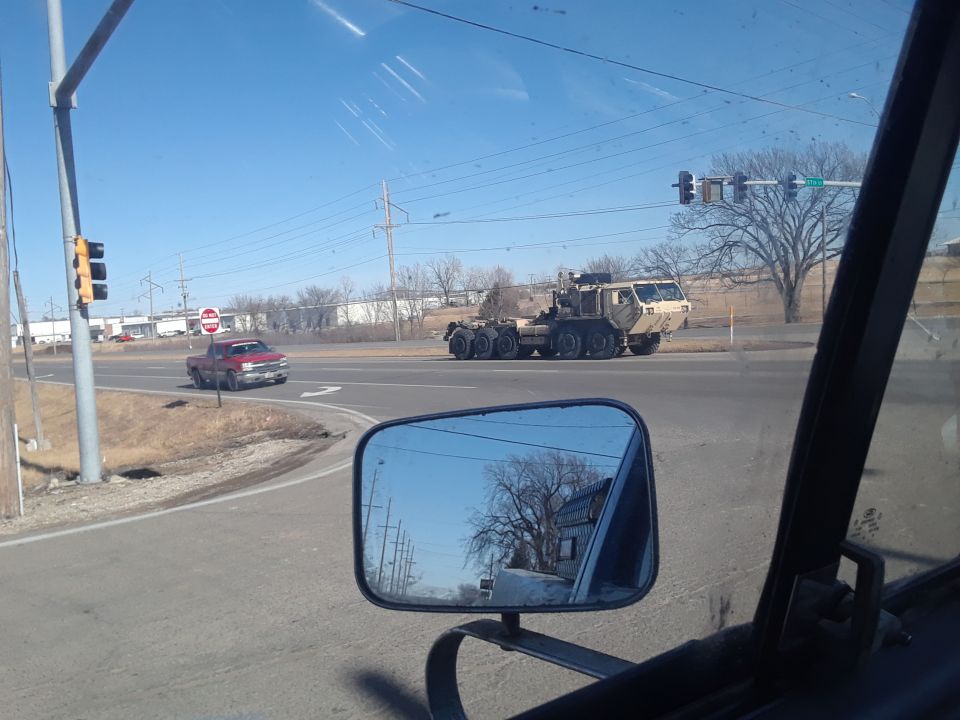 Tan military truck at intersection with red pickup truck, viewed from inside a vehicle.