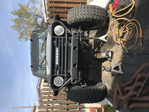 1987 Suzuki Samurai - Front view of a Suzuki Samurai, lifted on jack stands, with a spare tire and exhaust parts visible.