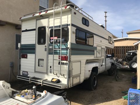 1997 Dodge 3500 - A camper is mounted on a dually truck, seen from the rear with a ladder on its side and a bicycle nearby.
