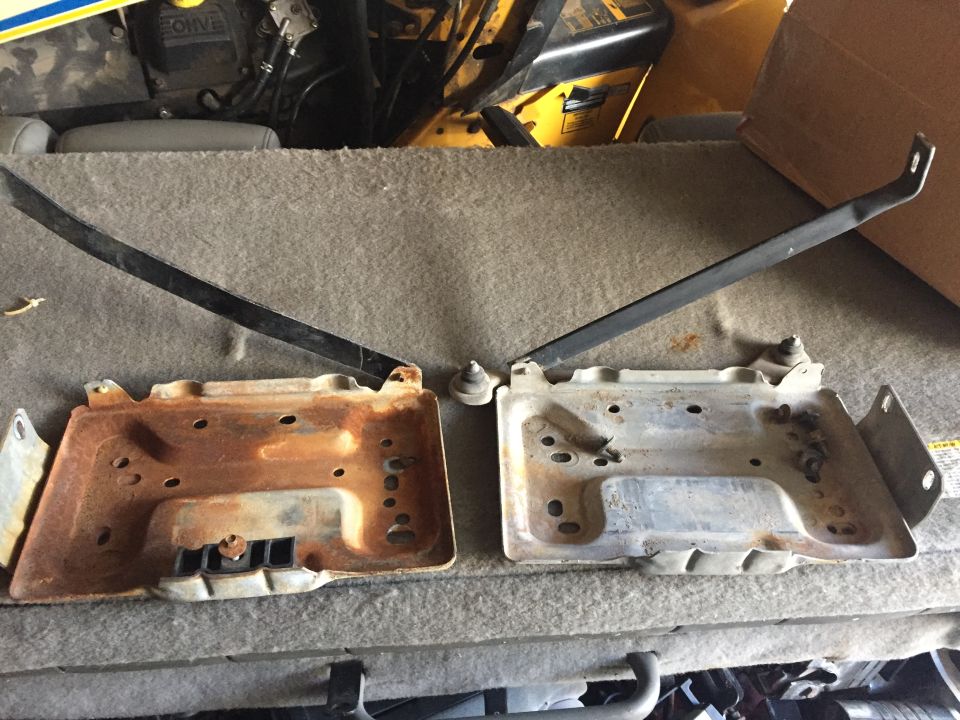 Two metal battery trays, one rusted, with black hold-down brackets. Parts are on a carpeted surface