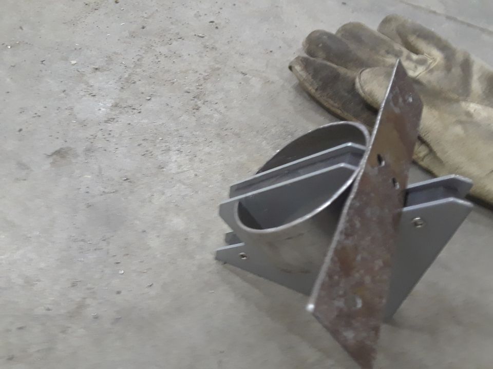 Metal bracket with a curved section and mounting holes, next to work gloves on a concrete floor
