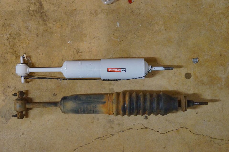 Two shock absorbers on a concrete floor; one is white with a red Motorcraft label, the other is black and rusty