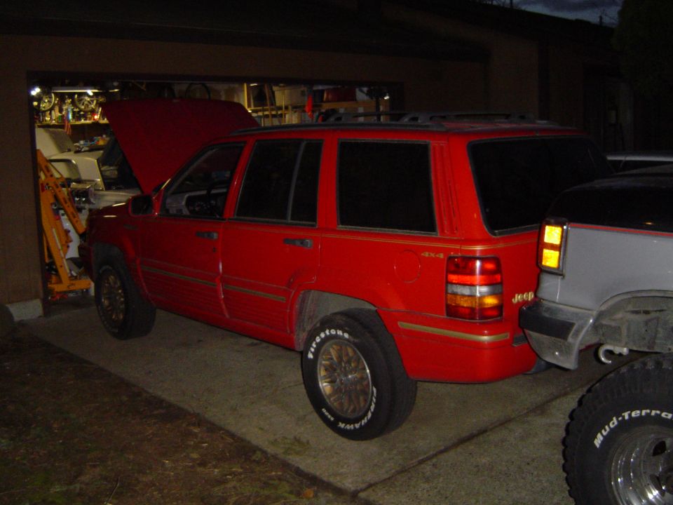 Red Jeep Grand Cherokee with hood up, parked at night by an open garage next to a silver truck