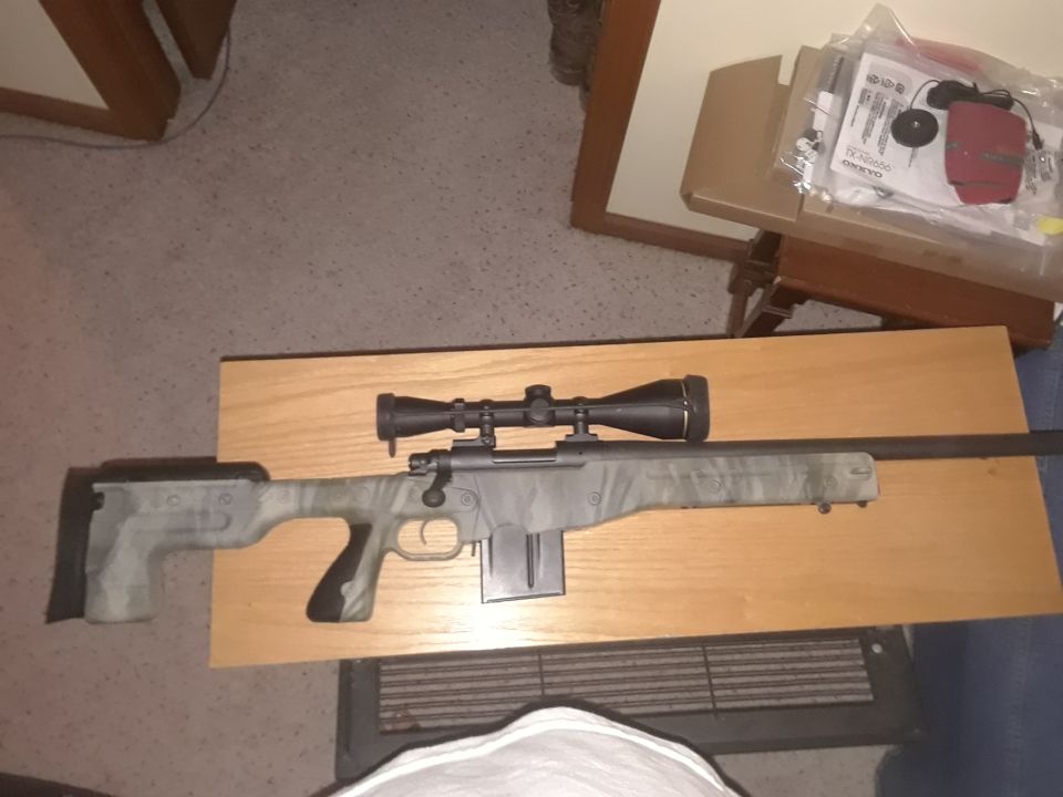 Camouflage rifle with scope on a wooden table, with electronics box in background.